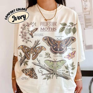 May include: An ivory t-shirt featuring a Field Guide to Moths graphic print. The design includes illustrations of a Hummingbird Clearwing, Cecropia, Rosy Maple, Atlas, Luna, and Garden Tiger moth with botanical elements and text labels.