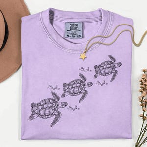 Puede incluir: Una camiseta Comfort Colors de color lila claro con una ilustraci&oacute;n negra de tres tortugas marinas nadando. Un collar dorado con un colgante de estrella est&aacute; sobre la camiseta. La etiqueta dice XL-TG-EG.