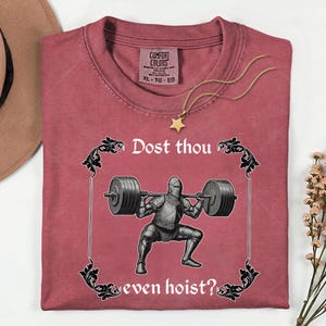 Op de afbeelding: Een bordeauxrood Comfort Colors T-shirt met een afbeelding van een ridder die gewichten heft, omlijst door sierlijke krullen. De tekst luidt "Dost thou even hoist?". Een gouden sterrenketting ligt op het shirt.