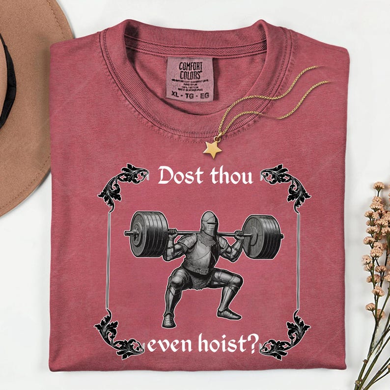 Op de afbeelding: Een bordeauxrood Comfort Colors T-shirt met een afbeelding van een ridder die gewichten heft, omlijst door sierlijke krullen. De tekst luidt "Dost thou even hoist?". Een gouden sterrenketting ligt op het shirt.