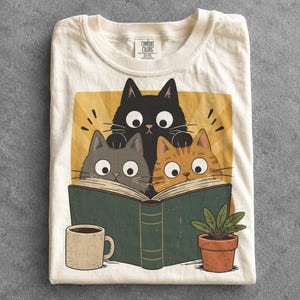 Könnte beinhalten: Ein cremefarbenes T-Shirt mit einer Grafik von drei Katzen – einer schwarzen, einer grauen und einer orangefarbenen –, die gemeinsam in einem großen grünen Buch lesen. Neben dem Buch stehen eine gesprenkelte beige Kaffeetasse und eine kleine Pflanze.