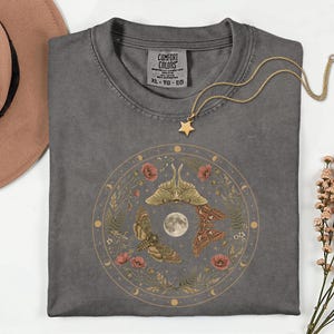Pode incluir: Uma t-shirt cinza-carvão com um gráfico circular de três traças, uma lua cheia e elementos florais. A t-shirt tem uma etiqueta Comfort Colors no colarinho. Um colar de corrente dourada com um pequeno pendente de estrela repousa no tecido.