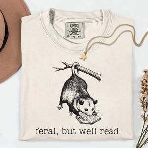 May include: Cream-colored t-shirt with a black and white illustration of an opossum reading a book, hanging from a branch. The text "feral, but well read" is printed below the opossum. A gold star pendant necklace is draped on the shirt.