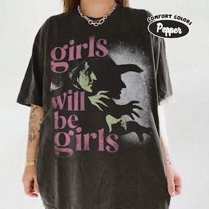 May include: A charcoal grey short-sleeve t-shirt featuring a graphic of a green-skinned witch in profile. The text "girls will be girls" is printed in pink lettering. A circular badge in the upper right corner reads "COMFORT COLORS Pepper".