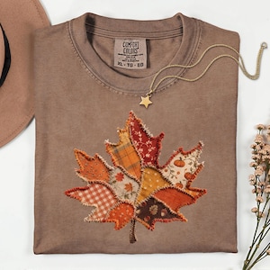Puede incluir: Una camiseta marrón con etiqueta Comfort Colors en talla XL, que presenta un gran diseño central de hoja de arce en patchwork hecho de varias telas estampadas en naranja, rojo y marrón con bordes deshilachados, junto a un collar con estrella dorada.
