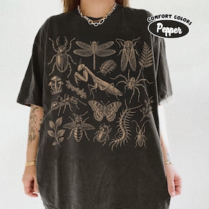 Op de afbeelding: Een antracietgrijs t-shirt met een gedetailleerde beige grafische print van diverse insecten, waaronder een bidsprinkhaan, libel, kever, spin, vlinder en duizendpoot, samen met botanische elementen en de tekst Comfort Colors Pepper.
