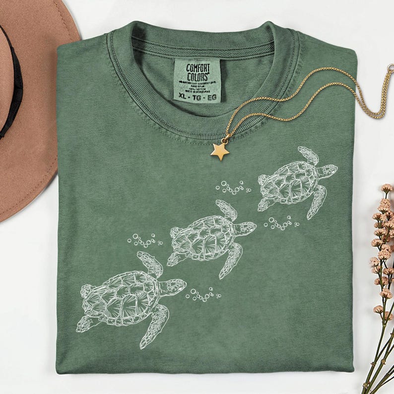 Puede incluir: Una camiseta verde salvia con un gr&aacute;fico blanco de tres tortugas marinas nadando. La camiseta tiene cuello redondo y mangas cortas. Un collar dorado con un colgante de estrella est&aacute; sobre la camiseta. La etiqueta dice "COMFORT COLORS XL-TG-EG".