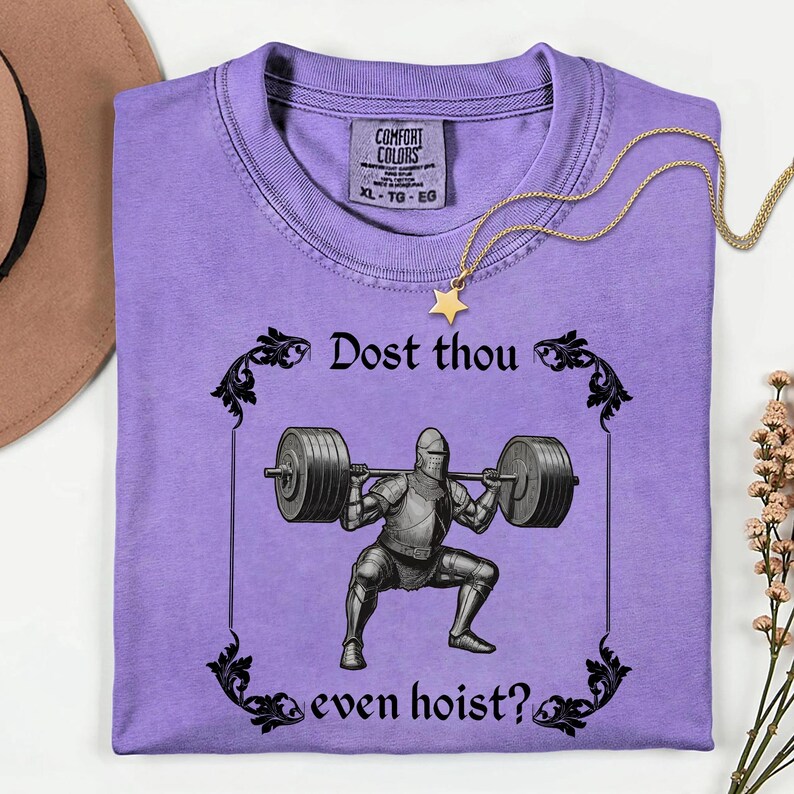 Op de afbeelding: Een lila Comfort Colors T-shirt met een ridder die gewichten heft, met de tekst "Dost thou even hoist?" in zwart. Een gouden ketting met een sterhanger ligt op het shirt.