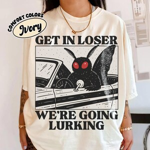 May include: An ivory short-sleeve t-shirt featuring a black graphic print of a moth-like creature driving a car. The text above reads GET IN LOSER and the text below reads WE'RE GOING LURKING. The design includes red eyes on the creature.