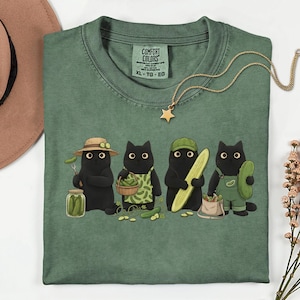 Puede incluir: Una camiseta verde salvia con un gráfico de cuatro gatos negros preparando pepinillos. Los gatos llevan sombreros y delantales, sosteniendo frascos, cestas y pepinos. La camiseta tiene una etiqueta Comfort Colors en el cuello y un collar de estrella.