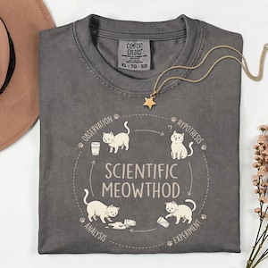 May include: A charcoal grey t-shirt featuring a circular graphic titled Scientific Meowthod. The design depicts four white cats illustrating steps of the scientific method: observation, hypothesis, experiment, and analysis, with paw prints and arrows.