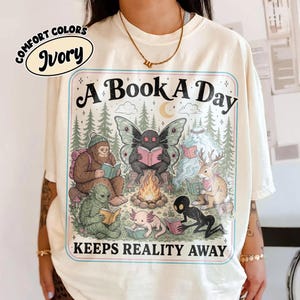 Può includere: Una t-shirt color avorio con una stampa grafica raffigurante una falena, un sasquatch, un cervo, un axolotl e altre creature che leggono attorno a un falò. Il testo recita A BOOK A DAY KEEPS REALITY AWAY. È visibile un'etichetta Comfort Colors Ivory.