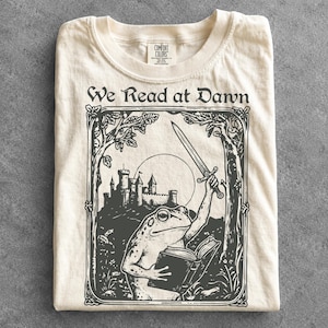 Puede incluir: Una camiseta de color crema con un estampado gráfico negro de una rana sosteniendo una espada y un libro abierto. La ilustración incluye un castillo y árboles dentro de un marco decorativo, con el texto "We Read at Dawn" sobre la imagen.