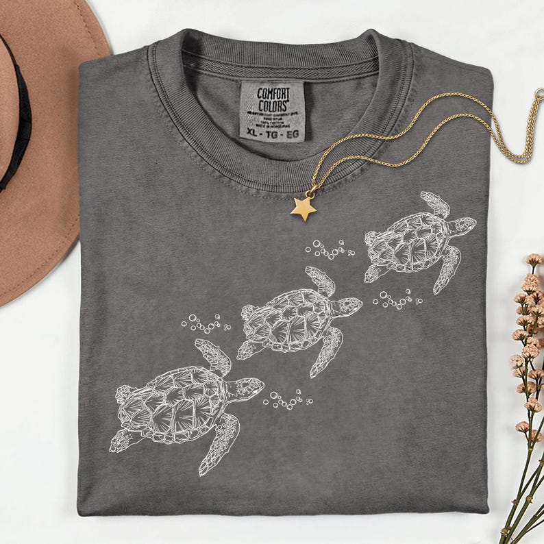 Puede incluir: Una camiseta Comfort Colors XL gris oscuro con un gr&aacute;fico blanco de tres tortugas marinas nadando. Tambi&eacute;n se ven un collar con colgante de estrella dorada y un sombrero marr&oacute;n. La camiseta tiene cuello redondo y mangas cortas.