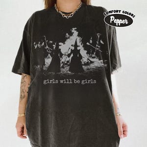 May include: A charcoal gray short-sleeve t-shirt featuring a graphic print of two figures in pointed hats standing before a bonfire. The text "girls will be girls" is printed in white below the graphic. A "COMFORT COLORS Pepper" label is visible.