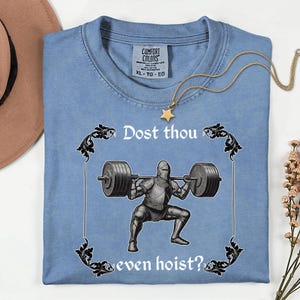 Op de afbeelding: Lichtblauw T-shirt met de tekst "Dost thou even hoist?" en een afbeelding van een ridder die gewichten heft. Het shirt heeft een Comfort Colors label en een gouden ketting met een sterhanger.
