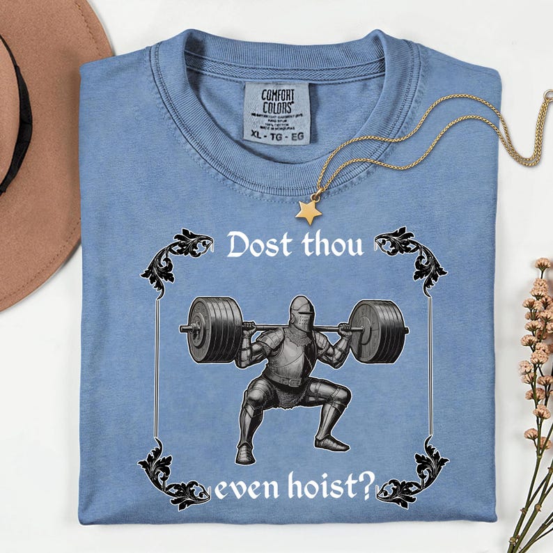 Op de afbeelding: Lichtblauw T-shirt met de tekst "Dost thou even hoist?" en een afbeelding van een ridder die gewichten heft. Het shirt heeft een Comfort Colors label en een gouden ketting met een sterhanger.