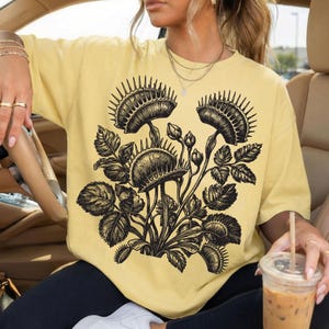 以下が含まれることがあります： 明るい黄色のTシャツ。黒と白の食虫植物のイラストが描かれています。デザインは、開いたハエトリグサ、葉、つぼみを示しています。ゆったりとしたフィット感のTシャツです。
