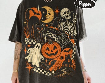 May include: A charcoal gray short-sleeve t-shirt featuring a graphic print with a black cat, a crescent moon, a skeleton, a ghost, a jack-o'-lantern, a witch hat, a spider web, bats, and a haunted house. The text Comfort Colors Pepper is visible.
