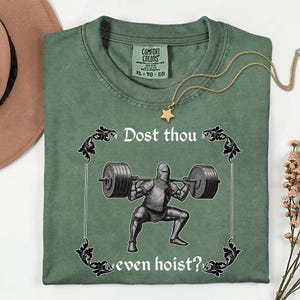 Op de afbeelding: Groen Comfort Colors T-shirt met de tekst "Dost thou even hoist?" en een afbeelding van een ridder die gewichten heft. Een gouden ketting met een sterhanger ligt op het shirt. Maat XL.
