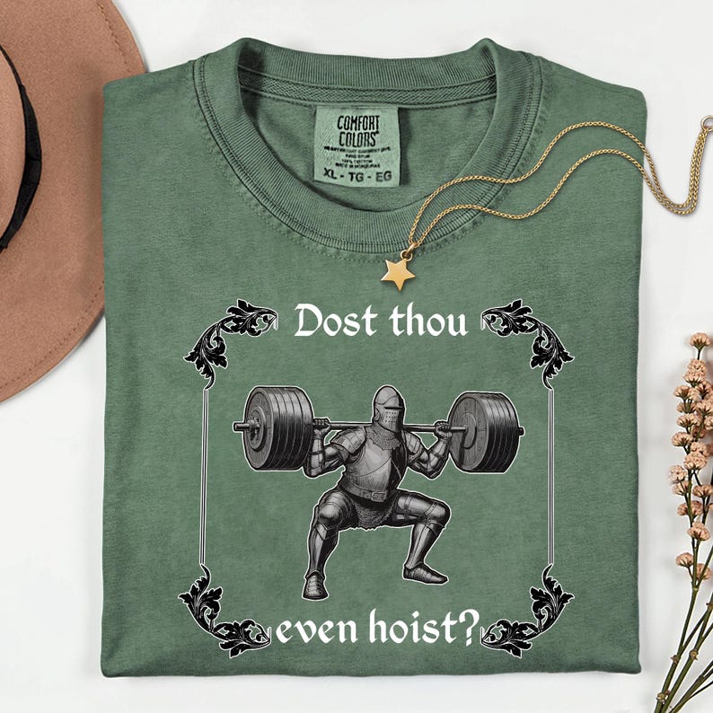 Op de afbeelding: Groen Comfort Colors T-shirt met de tekst "Dost thou even hoist?" en een afbeelding van een ridder die gewichten heft. Een gouden ketting met een sterhanger ligt op het shirt. Maat XL.