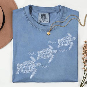 Puede incluir: Una camiseta Comfort Colors azul claro con un gráfico blanco de tres tortugas marinas nadando y burbujas. Un collar con colgante de estrella dorada descansa sobre la camisa. La talla de la camisa es XL-TG-EG.