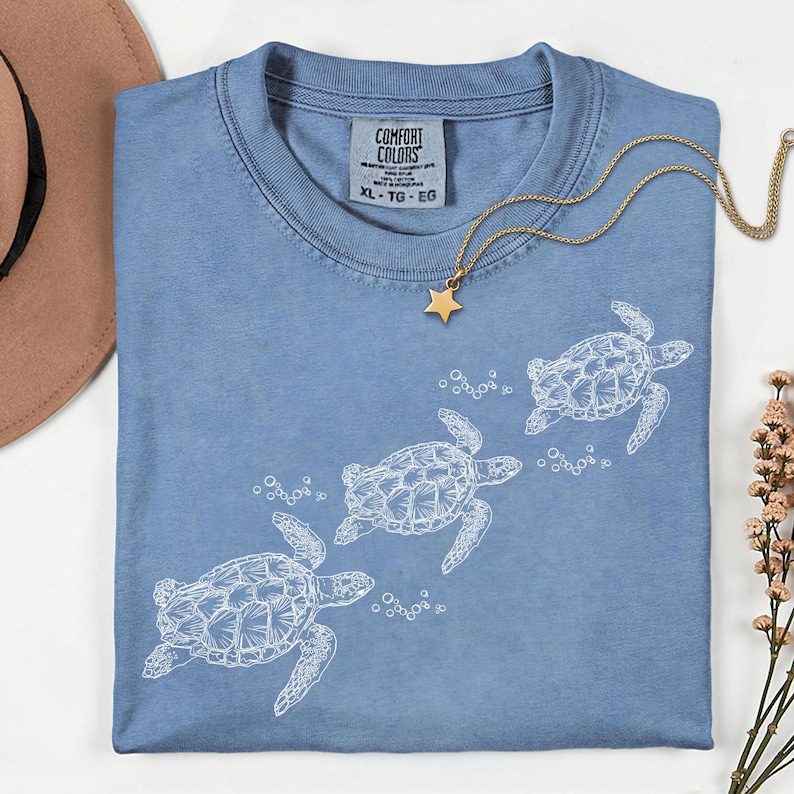 Puede incluir: Una camiseta Comfort Colors azul claro con un gr&aacute;fico blanco de tres tortugas marinas nadando y burbujas. Un collar con colgante de estrella dorada descansa sobre la camisa. La talla de la camisa es XL-TG-EG.