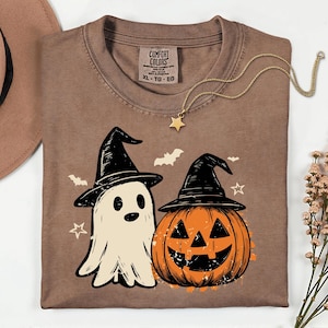 May include: A brown short-sleeve t-shirt featuring a graphic of a white ghost and an orange jack-o'-lantern, both wearing black witch hats. The design includes small white bats and stars. The shirt has a Comfort Colors label at the neck.