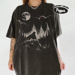 Puede incluir: Una camiseta color pimienta con un estampado gráfico de un paisaje forestal oscuro bajo una luna llena. El diseño incluye dos grandes criaturas similares a polillas con ojos rojos brillantes. El texto "COMFORT COLORS Pepper" aparece arriba.