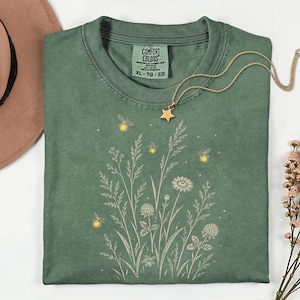 May include: A sage green t-shirt featuring a white line-art illustration of wildflowers and grasses with glowing yellow fireflies. The shirt has a Comfort Colors label at the neck, and a gold-toned necklace with a star charm rests on the fabric.