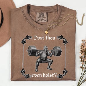 Op de afbeelding: Bruin Comfort Colors T-shirt met de tekst "Dost thou even hoist?" en een afbeelding van een ridder die gewichten heft. Een gouden sterrenketting is over het shirt gedrapeerd. Het shirt is op een wit oppervlak gevouwen.