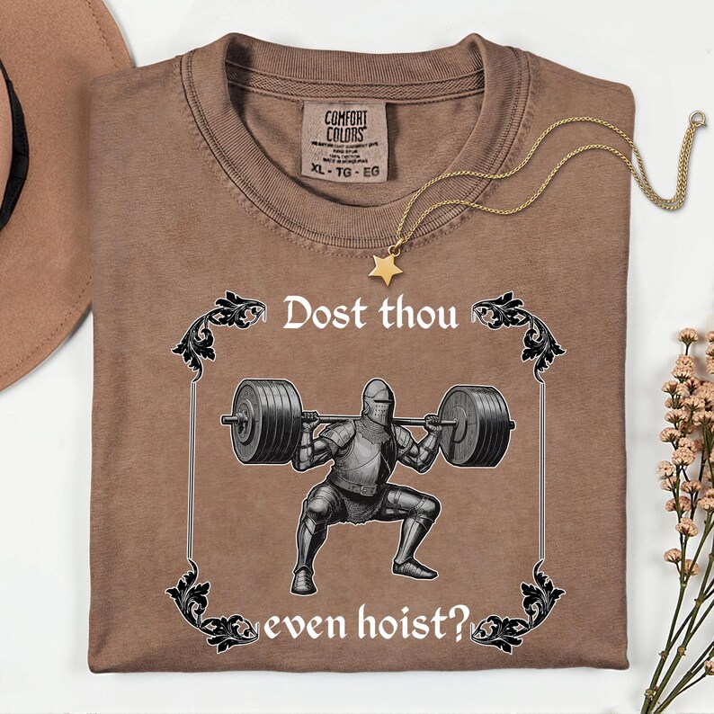 Op de afbeelding: Bruin Comfort Colors T-shirt met de tekst "Dost thou even hoist?" en een afbeelding van een ridder die gewichten heft. Een gouden sterrenketting is over het shirt gedrapeerd. Het shirt is op een wit oppervlak gevouwen.