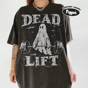Pode incluir: Uma t-shirt cinzenta de manga curta com um gráfico branco desgastado de um fantasma a levantar um halter. O texto "DEAD" aparece acima do fantasma e "LIFT" abaixo. Um emblema circular no canto superior direito diz "COMFORT COLORS Pepper".