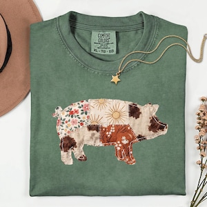 May include: A sage green short-sleeve t-shirt featuring a patchwork pig graphic. The pig design is composed of various fabric patterns including floral, sunburst, and brown cow print motifs. The shirt has a Comfort Colors label at the neck.