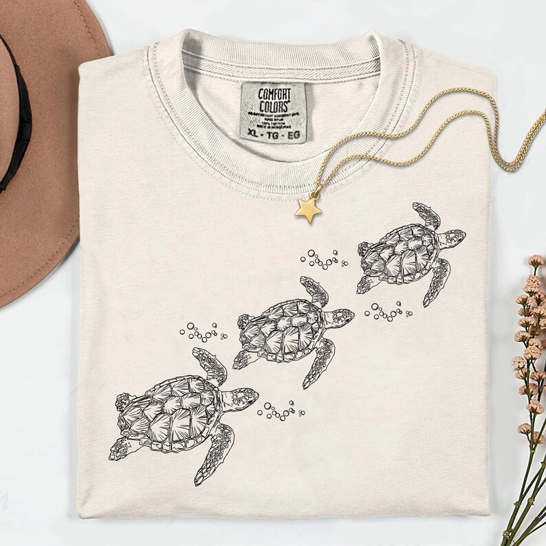 Puede incluir: Camiseta de color crema con un gr&aacute;fico de tres tortugas marinas nadando. La camiseta tiene cuello redondo y mangas cortas. Un collar dorado con un colgante de estrella est&aacute; drapeado sobre la camiseta. La etiqueta dice "COMFORT COLORS XL-TG-EG".