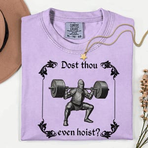 Op de afbeelding: Lichtpaars T-shirt met de tekst "Dost thou even hoist?" en een afbeelding van een ridder die gewichten heft. Het shirt heeft een ronde hals en korte mouwen. Een gouden ketting met een sterhanger ligt op het shirt.