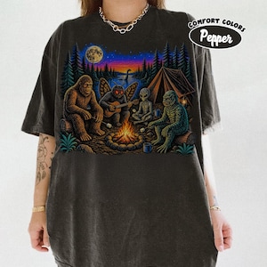 Może przedstawiać: Ciemnoszary t-shirt z nadrukiem przedstawiającym Wielką Stopę, ćmopodobne stworzenie grające na gitarze, kosmitę oraz potwora z bagien siedzących nocą przy ognisku. Grafika zawiera również namiot, pełnię księżyca oraz napis Comfort Colors Pepper.