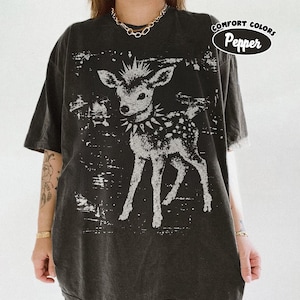 May include: A charcoal gray short-sleeve t-shirt featuring a distressed white graphic of a fawn wearing a spiked collar. The shirt has a faded, vintage-inspired texture. A circular badge in the upper right corner contains the text Comfort Colors Pepper.
