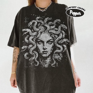 Puede incluir: Una camiseta de manga corta en color gris carbón con un gráfico grande y desgastado en blanco de Medusa con serpientes por cabello. Incluye una insignia circular en la esquina superior derecha con el texto Comfort Colors Pepper.