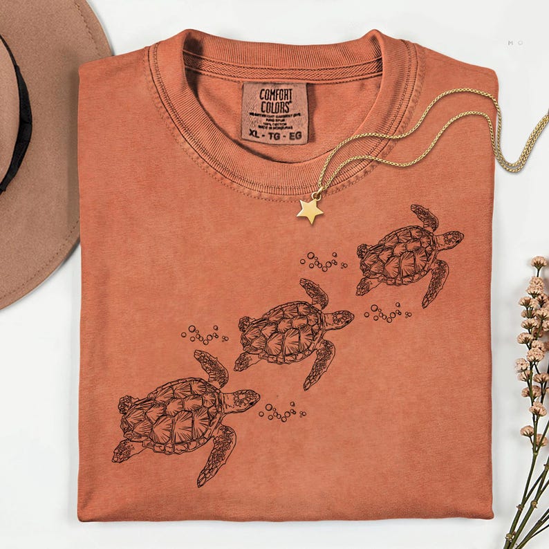 Puede incluir: Una camiseta Comfort Colors XL-TG-EG de color &oacute;xido con un dibujo lineal negro de tres tortugas marinas nadando. Un collar dorado con un colgante de estrella est&aacute; drapeado sobre la camisa. Un sombrero color canela y una ramita de flores est&aacute;n en el fondo.