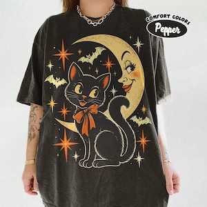 Puede incluir: Una camiseta gris carbón con un gráfico de un gato negro con un lazo naranja, una luna creciente con cara, murciélagos y estrellas naranjas. El texto COMFORT COLORS Pepper está impreso en la esquina superior derecha del diseño.