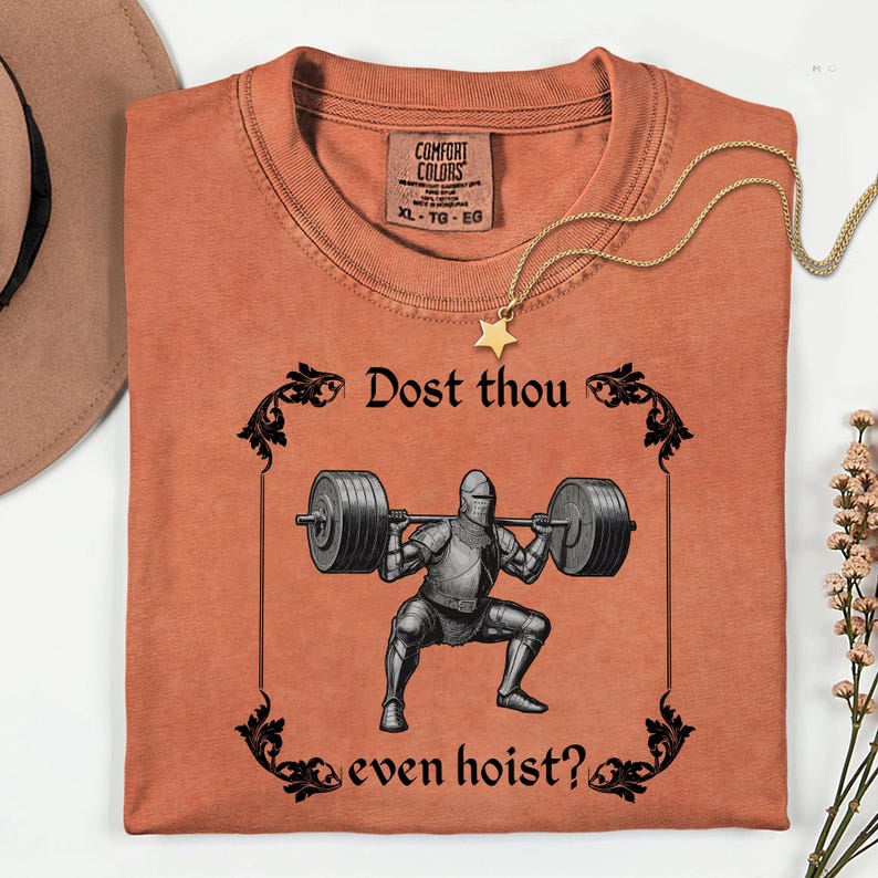 Op de afbeelding: Oranje Comfort Colors T-shirt met een zwarte afbeelding van een ridder die gewichten heft, omlijst door decoratieve krullen. De tekst luidt "Dost thou even hoist?" Een gouden sterhangerketting en een hoed zijn zichtbaar.