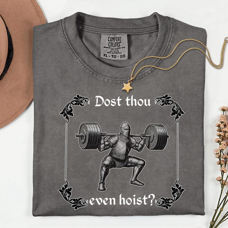Op de afbeelding: Donkergrijs t-shirt met de tekst "Dost thou even hoist?" en een illustratie van een ridder die gewichten heft. Het shirt heeft een Comfort Colors label en een gouden sterrenketting.