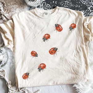 May include: A cream-colored short-sleeve t-shirt featuring a scattered pattern of seven red ladybugs with black spots. The shirt has a crew neckline and a relaxed fit, with the ladybugs distributed across the front of the fabric in various sizes.