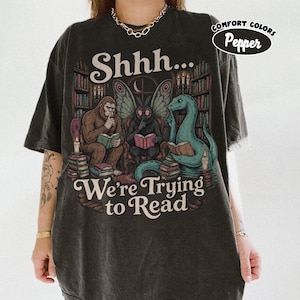 Può includere: Una t-shirt grigio antracite con una grafica di Bigfoot, una creatura simile a una falena e un mostro marino che leggono in una biblioteca. Il testo recita Shhh... We're Trying to Read. Un'etichetta circolare indica Comfort Colors Pepper.
