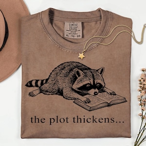 May include: Brown t-shirt with a raccoon reading a book, and the text "the plot thickens...". The shirt has a crew neck and a tag that says "Comfort Colors". A gold star pendant necklace is draped across the shirt.