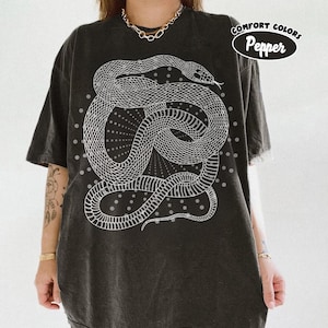 Puede incluir: Una camiseta de manga corta gris carbón con un gran gráfico de líneas blancas de una serpiente enroscada rodeada por un patrón de rayos de sol punteados. La camiseta incluye un logotipo circular con el texto Comfort Colors Pepper.