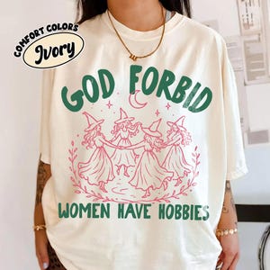 Puede incluir: Una camiseta de manga corta color marfil con un gráfico verde oscuro que incluye el texto "GOD FORBID WOMEN HAVE HOBBIES" y una ilustración central de cuatro figuras con sombreros puntiagudos tomadas de la mano en círculo, con detalles en rosa.