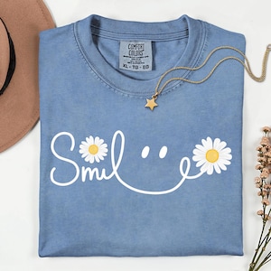 Puede incluir: Camiseta azul claro Comfort Colors con la palabra "Smile" en escritura blanca, con detalles de margaritas. Un collar dorado con un colgante de estrella descansa sobre la camiseta. Talla XL.