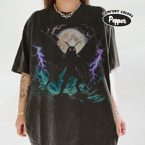 Może przedstawiać: Ciemnoszary t-shirt z krótkim rękawem z grafiką przedstawiającą ćmopodobne stworzenie ze świecącymi na czerwono oczami na tle pełni księżyca. Wzór zawiera fioletowe i turkusowe błyskawice. Naklejka w rogu zawiera napis COMFORT COLORS Pepper.